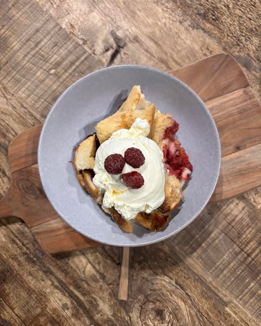 A warm pudding with whipped cream and fresh raspberries on a timber board